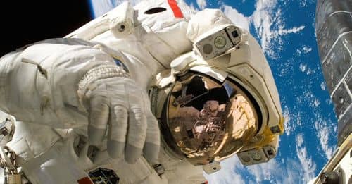 This picture shows an american astronaut in his space and extravehicular activity suite working outside of a spacecraft. In the background parts of a space shuttle are visible. In the far background of the picture planet earth with it's blue color and whi