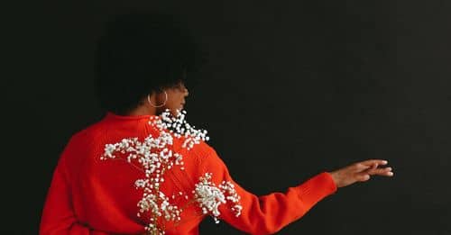 Back view of modern African American model wearing vivid red sweater with white bottom holding baby breath flower behind back on black background