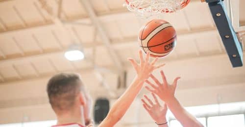 From below adult men in red and white sportswear catching ball from basket while playing basketball on sports arena in light contemporary sports club