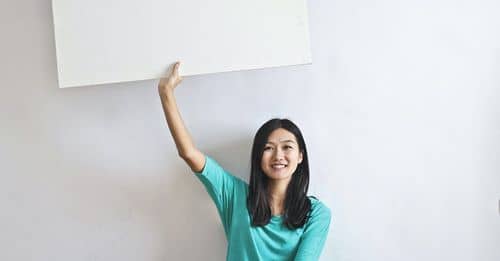 Cheerful Asian woman sitting cross legged on floor against white wall in empty apartment and showing white blank banner