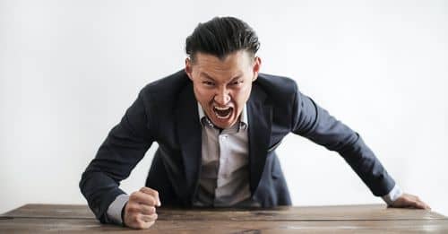 Expressive angry businessman in formal suit looking at camera and screaming with madness while hitting desk with fist