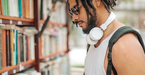 Confident adult Hispanic male in sunglasses and casual clothes with backpack and headphones reading book near bookshelf while making choice in stall on street