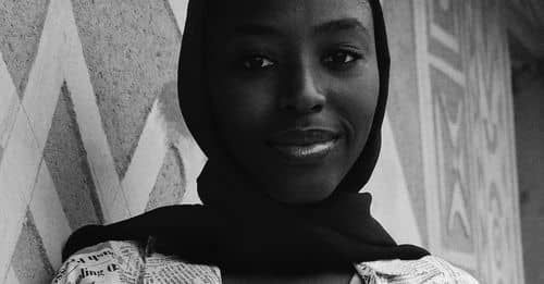 Black and white low angle of young smiling African American female in shawl on head and t shirt with text pattern standing leaned on concrete wall and looking at camera