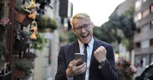 Excited young man in formal wear and eyeglasses clenching fist and rejoicing victory while using mobile phone in modern city street