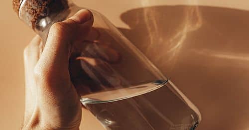 Crop anonymous male holding transparent glass bottle of water in hand against beige wall with shades during sunny day