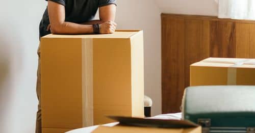 Thoughtful ethnic man standing in new house leaning on unpacked carton box