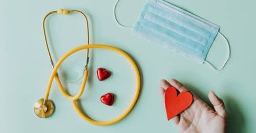 Top view of crop anonymous person hand with red paper heart on table with stethoscope and medical mask for coronavirus prevention