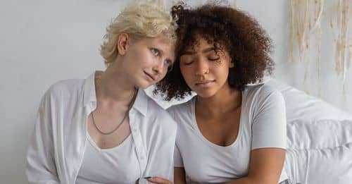 African American lady with closed eyes cuddling female friend while sitting on bed with opened suitcase during packing things in cozy room