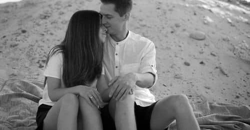 Black and white of young content man caressing female beloved while resting on hillside under cloudy sky