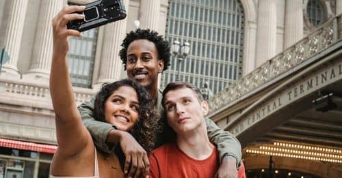 Positive multiethnic friends taking selfie on modern smartphone near railway station building