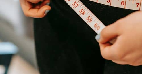 From above of crop anonymous plump female using measuring tape around hips in gym