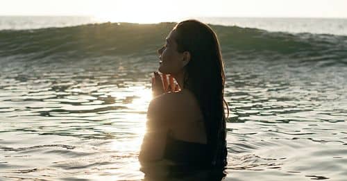 Side view of tranquil young female tourist with long dark hair standing in waving ocean with closed eyes and enjoying summer sunset