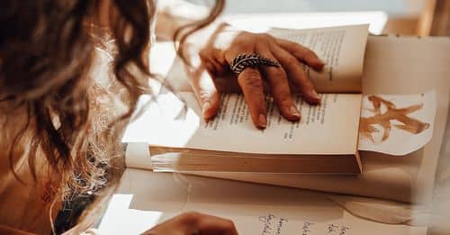 Crop anonymous female with dark hair sitting near window at retro writing cabinet while taking notes with feather in vintage store