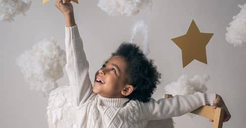 Cheerful African American child with angel wings touching decorative star while looking up from chair on light background