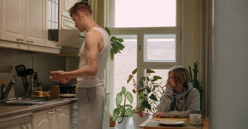 A Young Boy in Gray Sweater Sitting Near the Table while Looking at His Brother Standing Near the Kitchen Cabinets