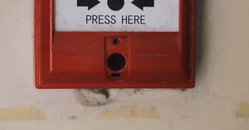 Manual emergency red fire alarm system box with push button placed on white shabby wall in light room of building