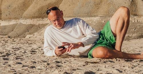 Man in White Sweater Lying on Sand and Using Mobile Phone