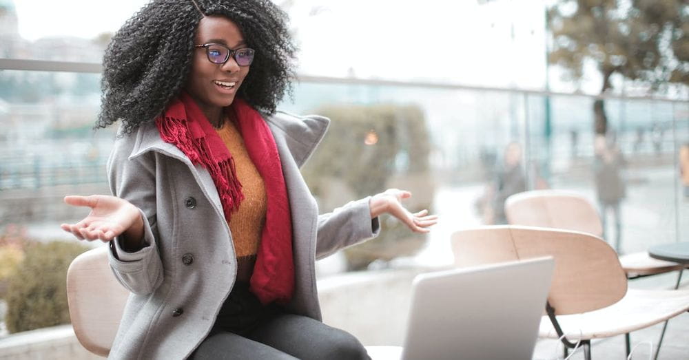 Happy excited African American female laughing and gesticulating while having video calling on laptop and sitting at modern cafe