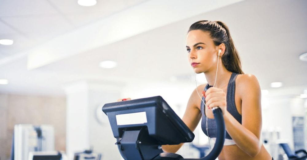 Serious fit woman in earphones and activewear listening to music and running on treadmill in light contemporary sports center