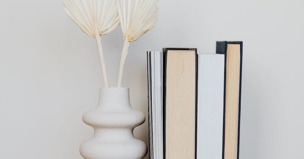 White original vase on wooden round table along with stack of assorted books in front of white wall