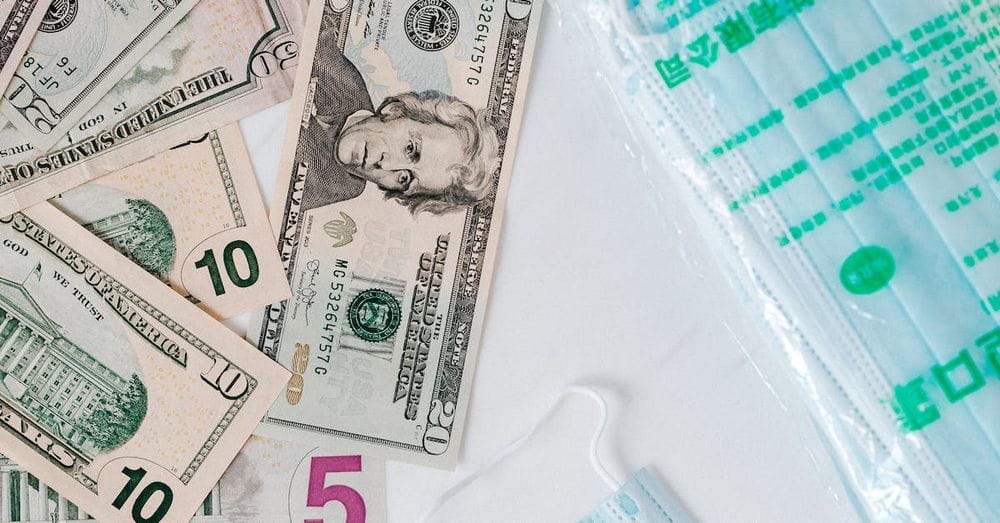 Top view composition of disposable medical masks placed on white table near scattered randomly various paper banknotes of American dollars