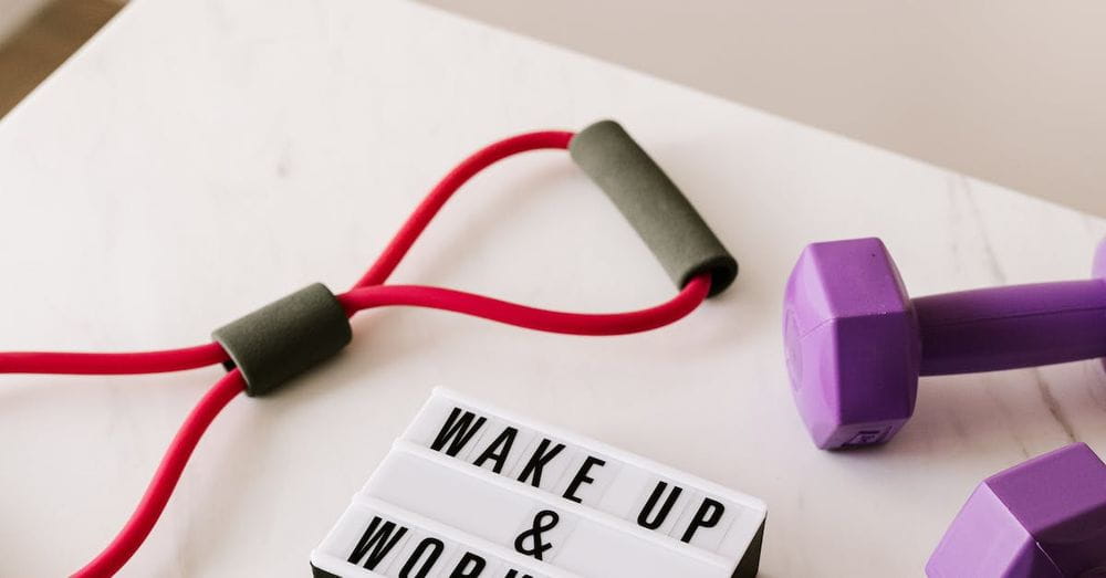 From above composition of dumbbells and massage double ball and tape and tubular expanders surrounding light box with wake up and workout words placed on white surface of table