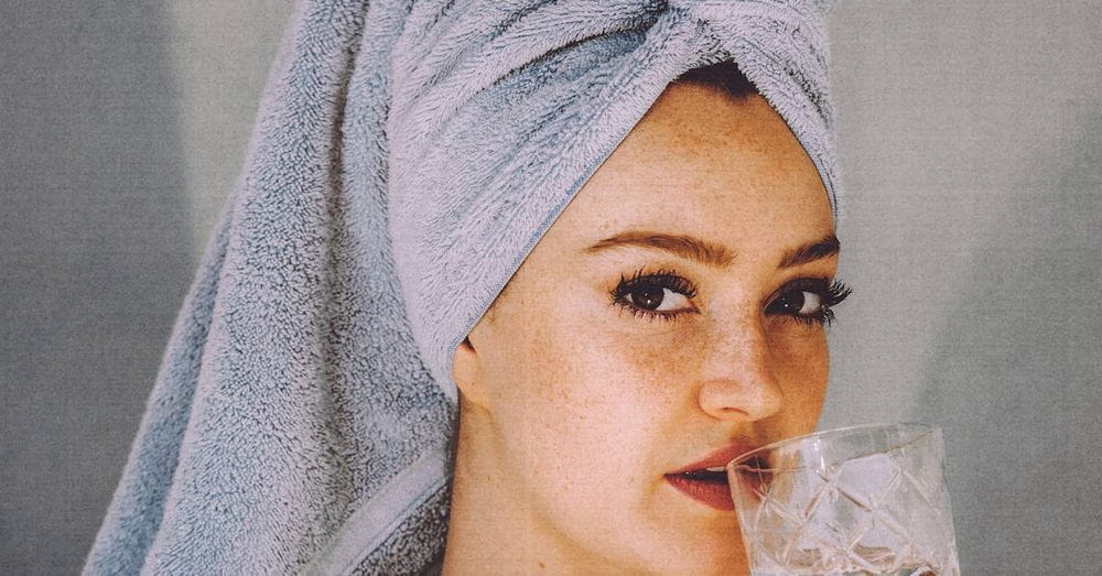 Young woman with hair wrapped with towel and bared shoulders drinking water and looking at camera