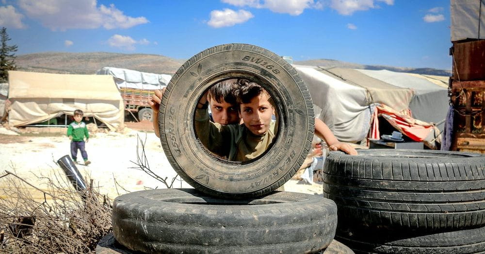 Pensive boys looking through big black wheel of vehicle under vibrant blue sky with clouds in encampment