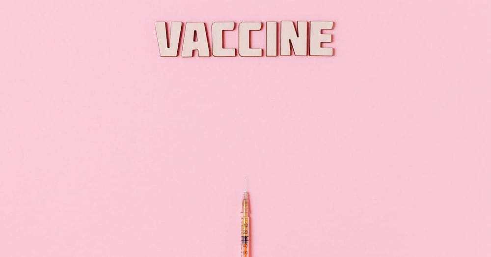 Vaccine Text and A Person Wearing Latex Glove While Holding a Syringe on Pink Background