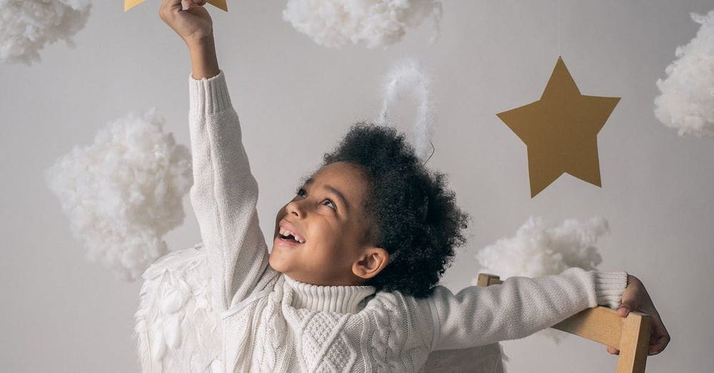 Cheerful African American child with angel wings touching decorative star while looking up from chair on light background