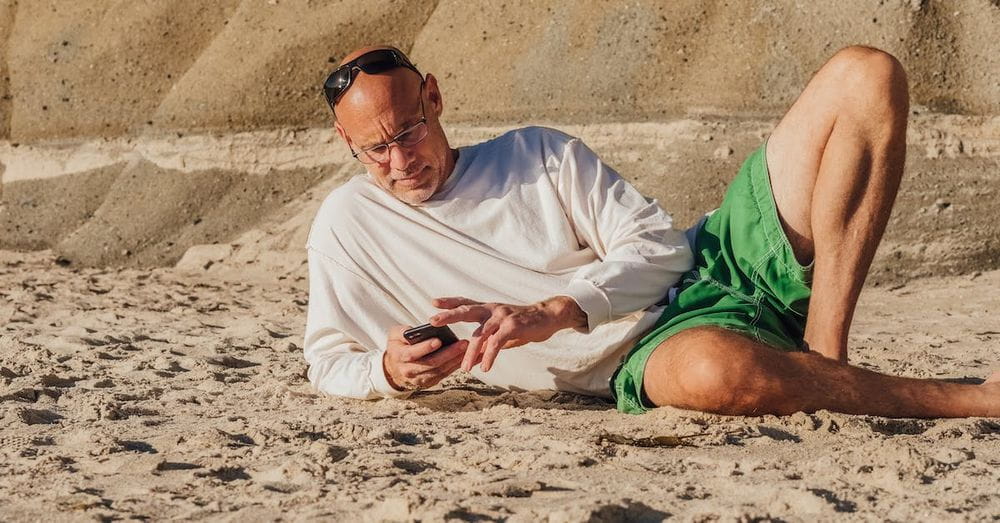 Man in White Sweater Lying on Sand and Using Mobile Phone