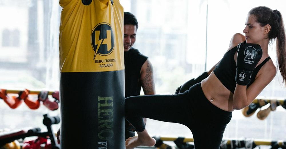 Strong female kickboxer in gloves kicking heavy punching bag near personal Latin American instructor during intense training in modern gym